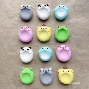 Puede incluir: Una colección de doce figuras de animales redondeadas y coloridas. Las figuras incluyen un panda, un koala, una rana, un cerdo y otras criaturas en varios colores como azul, rosa y amarillo. Cada una tiene una expresión facial y un diseño únicos.