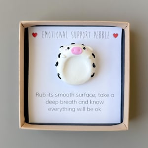 May include: An emotional support pebble in a box. The white pebble is shaped like a pig with black spots and a pink snout. The box has text that reads "EMOTIONAL SUPPORT PEBBLE" and "Rub its smooth surface, take a deep breath and know everything will be ok."
