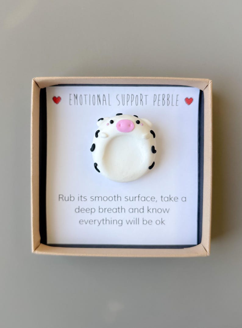 May include: An emotional support pebble in a box. The white pebble is shaped like a pig with black spots and a pink snout. The box has text that reads "EMOTIONAL SUPPORT PEBBLE" and "Rub its smooth surface, take a deep breath and know everything will be ok."