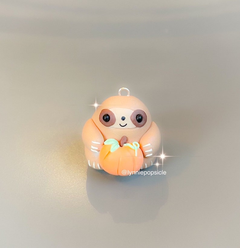 May include: A small, brown sloth figurine holding an orange pumpkin. The sloth has a friendly face and is made of polymer clay. The figurine is a charming autumn decoration.