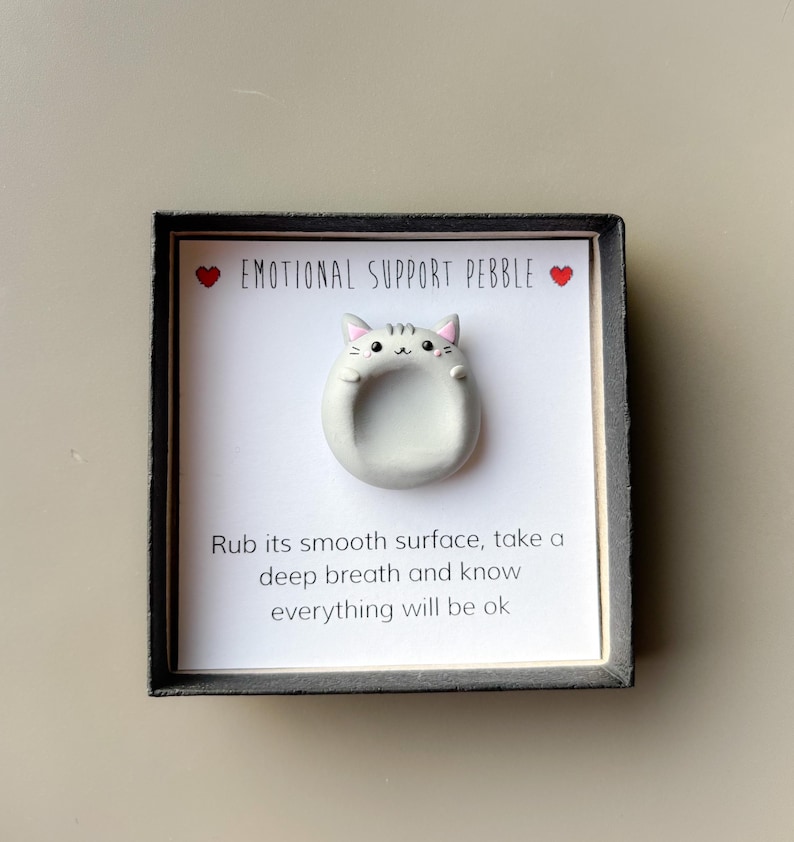 May include: A gray, cat-shaped emotional support pebble on a white card, framed in black. The card reads "EMOTIONAL SUPPORT PEBBLE" with heart symbols. Below, text encourages the user to rub the smooth surface and breathe.
