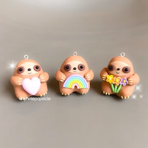 May include: Three brown sloth ornaments, each holding a different item. The first sloth is holding a pink heart, the second is holding a rainbow, and the third is holding a bouquet of yellow, orange, and purple flowers.