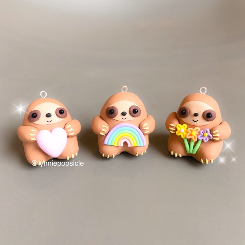 May include: Three brown sloth ornaments, each holding a different item. The first sloth is holding a pink heart, the second is holding a rainbow, and the third is holding a bouquet of yellow, orange, and purple flowers.