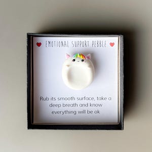 May include: A white, oval-shaped "Emotional Support Pebble" with a unicorn face and rainbow mane, presented in a black box. The text reads "Rub its smooth surface, take a deep breath and know everything will be ok."