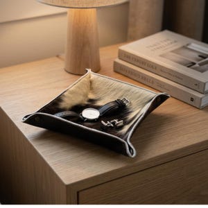May include: A square valet tray featuring a textured cowhide pattern in shades of black, white, and cream. The tray has raised, snap-button corners and contains a black leather-strap wristwatch with a white face and a pair of silver-toned cufflinks.