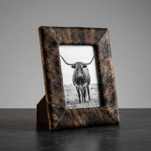 May include: A rectangular picture frame featuring a textured brown and black cowhide finish. The frame contains a black and white photograph of a longhorn steer standing in a field. The frame is supported by a stand on the back side.