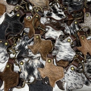 May include: A pile of brown and white cowhide keychains shaped like the state of Texas. The keychains have a brown leather strap and a metal ring.