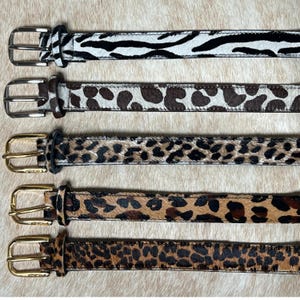 May include: A collection of five animal print belts. The belts feature patterns such as zebra stripes, leopard spots, and cheetah print. Each belt has a silver or gold-colored buckle. The belts are displayed on a light-colored, textured surface.