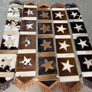 May include: Six cowhide rugs with fringe edges. Each rug has a brown, white, and black patchwork design with a large white star in the center.