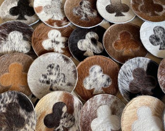 May include: A set of 14 round cowhide coasters with a black club symbol in the center of each coaster. The coasters are made of different colors and patterns of cowhide.