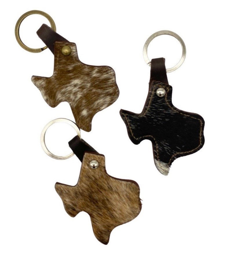 May include: Three leather keychains shaped like the state of Texas. One keychain is brown with white spots, one is black with white spots, and one is brown with white and black spots.