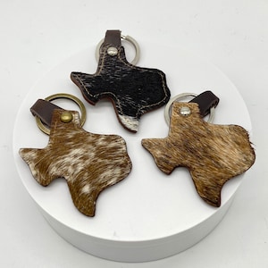 May include: Three leather keychains shaped like the state of Texas. One keychain is black and white, one is brown and white, and one is light brown and white.