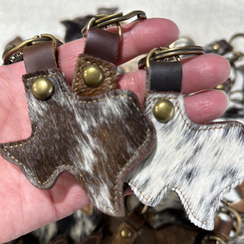 May include: Two leather keychains shaped like the state of Texas. The keychains are made of brown and white cowhide with a brown leather strap and a metal key ring.