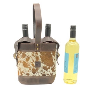 May include: A brown leather and cowhide wine carrier with a single handle, designed to hold two bottles. The carrier features a brown leather base and top trim, with a central cowhide panel. A separate bottle of white wine stands next to the carrier.