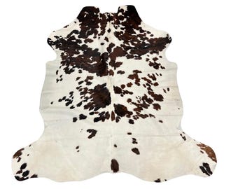 May include: A cowhide rug featuring an irregular, organic shape with a natural pattern of dark brown and black patches distributed across a creamy white background. The hide displays a smooth, flat texture with distinct, asymmetrical edges.
