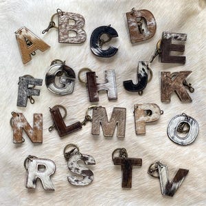 May include: A set of 26 cowhide leather keychains, each featuring a different letter of the alphabet. The keychains are brown and white with a spotted pattern.