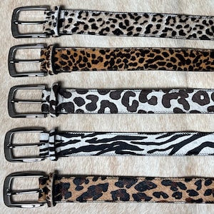May include: Five leather belts with silver-toned buckles. The belts feature animal print patterns, including leopard, cheetah, and zebra designs. The belts are displayed on a textured, light brown surface.