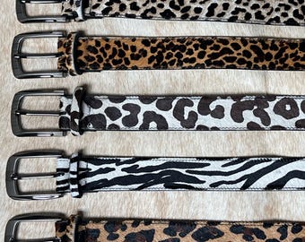 May include: Five leather belts with silver-toned buckles. The belts feature animal print patterns, including leopard, cheetah, and zebra designs. The belts are displayed on a textured, light brown surface.