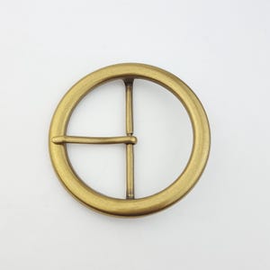 Puede incluir: Hebilla de cinturón redonda en acabado latón antiguo. La hebilla presenta un diseño clásico con un marco redondo y una barra transversal. El metal tiene una superficie texturizada y un cálido tono dorado. Adecuada para cinturones de varios anchos.