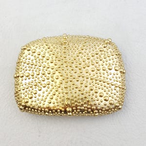 Pode incluir: Uma fivela de cinto de metal dourado com uma superfície texturizada. A fivela é coberta por pequenos pontos redondos em relevo.
