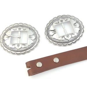 May include: Two silver-colored oval belt buckles with a floral and arrow design. The buckles have a square center and a decorative edge. A brown leather strap with two silver rivets is also visible.