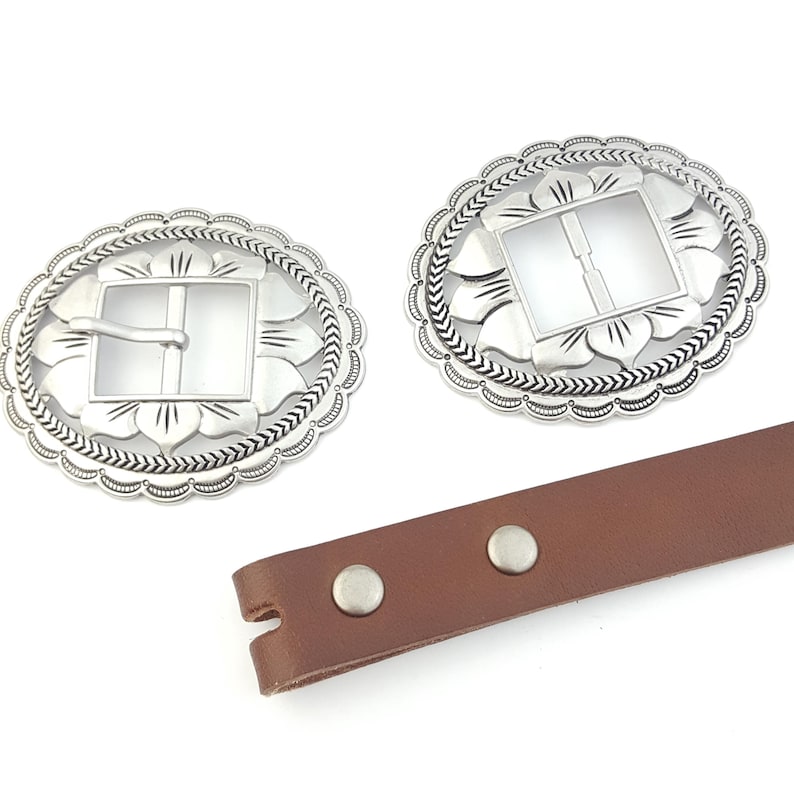 May include: Two silver-colored oval belt buckles with a floral and arrow design. The buckles have a square center and a decorative edge. A brown leather strap with two silver rivets is also visible.