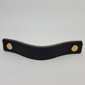 Puede incluir: Tirador de cajón de cuero negro con diseño curvo. El tirador se asegura con dos remaches dorados. El cuero tiene un aspecto texturizado y el diseño general es simple y moderno. Adecuado para armarios y cajones.