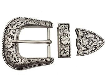 May include: Silver-toned western belt buckle set with intricate floral and scrollwork designs. The set includes a buckle, belt loop, and tip, all with a detailed, engraved pattern. The metal has a slightly aged appearance.