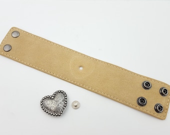 May include: A tan leather bracelet with silver metal accents. The bracelet has a heart-shaped charm with an intricate design.