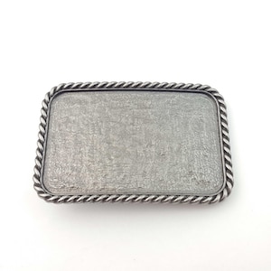 Puede incluir: Una hebilla de cinturón de plata con un borde de diseño de cuerda. La hebilla está en blanco y está lista para ser personalizada.