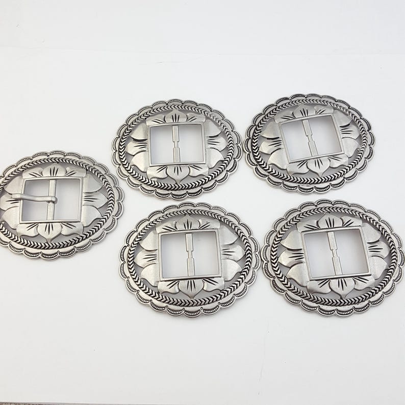 May include: Five silver-colored oval belt buckles with a floral design around the edges. Each buckle has a square opening for the belt strap. The buckles are made of metal and have a vintage or Western aesthetic.