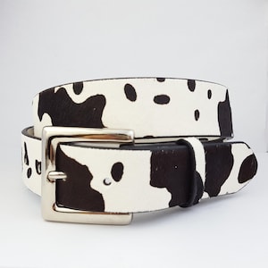 Pode incluir: Um cinto com padrão de pele de vaca e fivela prateada. O cinto apresenta um design manchado em preto e branco. A fivela é quadrada e tem um acabamento brilhante. É um acessório de moda.