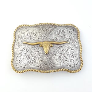 May include: Silver-toned rectangular belt buckle with a gold-colored longhorn steer head in the center. The buckle has an ornate floral design and a gold rope-like border. A Western-themed accessory.