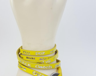 May include: Yellow leather wrap bracelet with silver lettering. The lettering reads "Yell Skip Scoot Run We Up Get Up Catch Over Come".