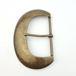 Puede incluir: Hebilla de cinturón de latón antiguo con forma ovalada redondeada. La hebilla tiene un diseño clásico con una barra horizontal y dos barras verticales para la correa del cinturón. El metal tiene un acabado ligeramente texturizado.