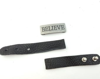 May include: A black leather bracelet with two silver metal snaps and a silver metal charm that says "BELIEVE".