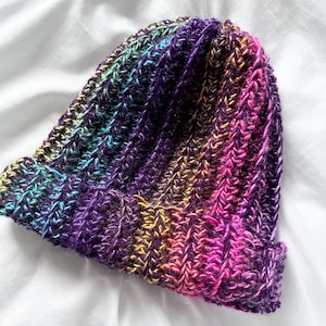 May include: A colourful crocheted beanie hat with a purple, pink, yellow and blue colour scheme. The hat is made with a textured stitch and has a slouchy fit.