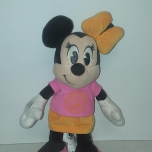 May include: A plush Minnie Mouse doll with a pink shirt, orange skirt, and a yellow bow. The doll has black ears, a black nose, and pink shoes. The doll is standing upright.