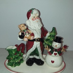 May include: A ceramic Christmas tea set featuring Santa Claus holding a teddy bear, a snowman with a red heart, and a green Christmas tree. The set includes a creamer, sugar bowl, and a tray.