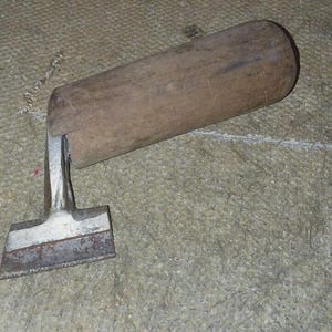 May include: A vintage woodworking chisel with a wooden handle and a metal blade. The handle is cylindrical and made of light brown wood. The metal blade is rectangular and shows signs of wear.