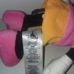 May include: A plush Daisy Duck toy, featuring a yellow head, pink bow, and pink and black feet. The toy has a white hand and a pink dress. A Disney tag is visible.