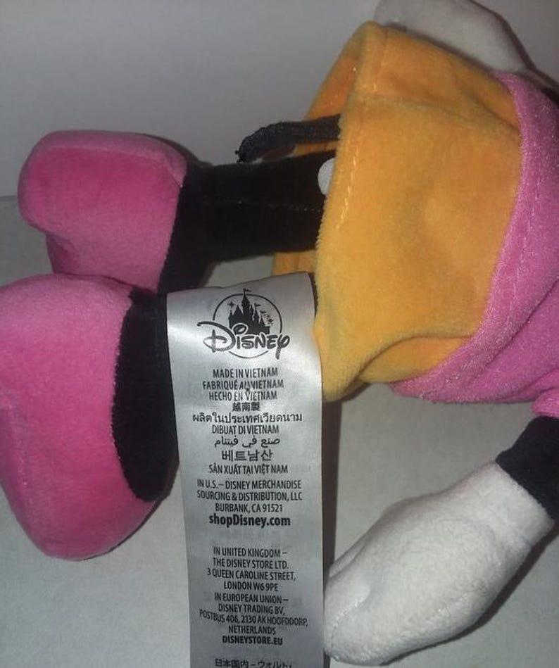 May include: A plush Daisy Duck toy, featuring a yellow head, pink bow, and pink and black feet. The toy has a white hand and a pink dress. A Disney tag is visible.