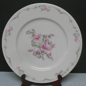 May include: White ceramic dinner plate with a gray and pink floral design. The plate has a scalloped edge and a gray and white ribbon design around the rim.