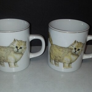 May include: Two white ceramic mugs with a gold rim and handle. Each mug features a detailed illustration of a fluffy, light-brown kitten. The kitten has black eyes and is standing upright. The mugs are ideal for coffee or tea.