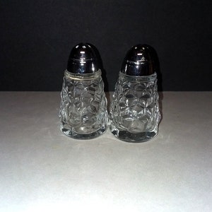 May include: Two clear glass salt and pepper shakers with a textured, diamond-like pattern. Each shaker has a shiny, silver-colored metal lid with small holes for dispensing. The shakers are displayed on a white surface against a dark background.