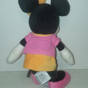 May include: A plush Minnie Mouse doll, viewed from the back, with black ears and head, wearing a pink top and yellow skirt. The doll has white gloves and pink shoes. A yellow bow is on her head.