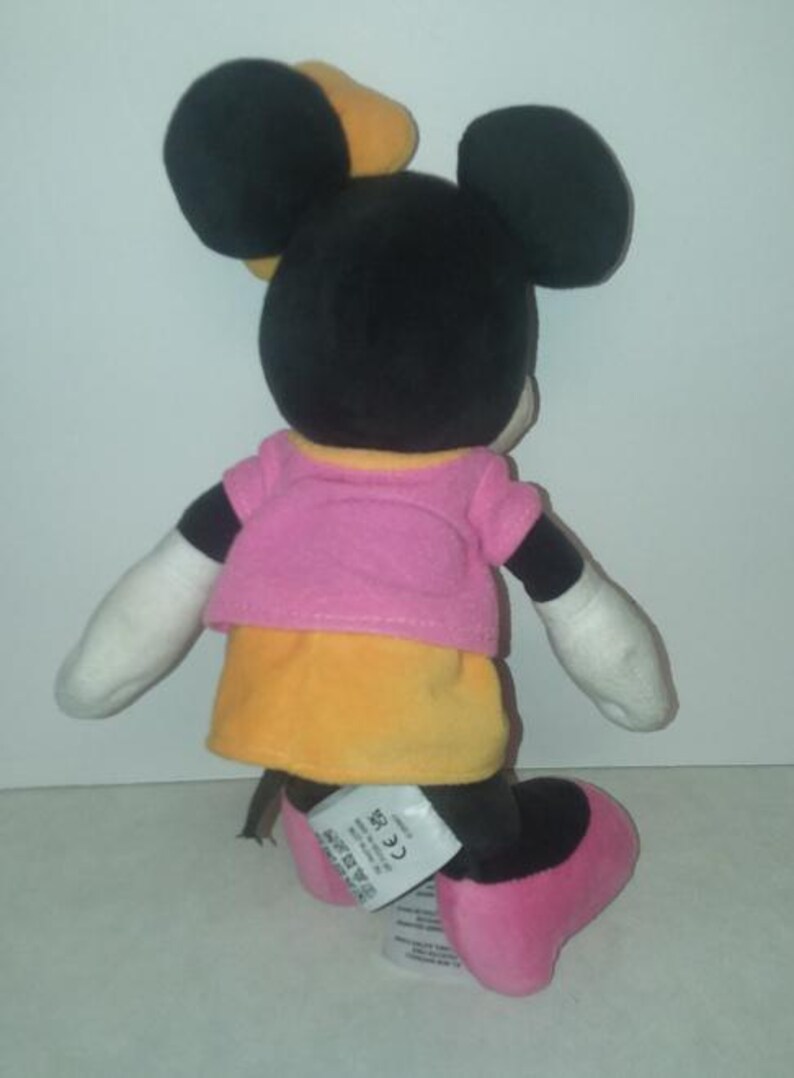 May include: A plush Minnie Mouse doll, viewed from the back, with black ears and head, wearing a pink top and yellow skirt. The doll has white gloves and pink shoes. A yellow bow is on her head.