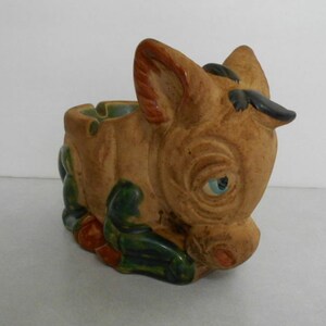 May include: A ceramic donkey ashtray with green, brown, and orange glaze. The donkey has a blue eye and a black nose.