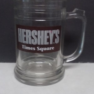 May include: A clear glass mug with a brown and white Hershey's Times Square logo.