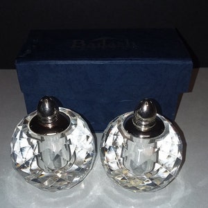 May include: Two clear, faceted crystal salt and pepper shakers with silver-colored tops. The shakers are spherical and sit in front of a dark blue box. The box has the word "Dadash" printed on it.